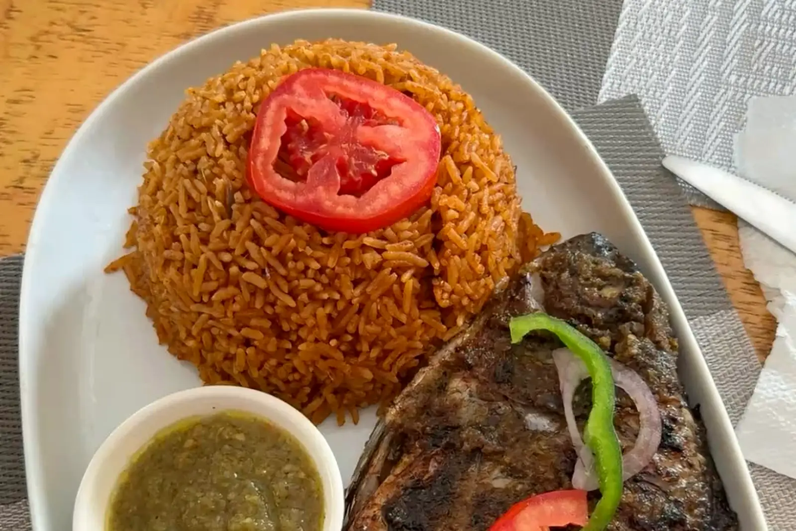 Red-orange jollof rice with whole grilled fish, green pepper, onion, tomato and a small bowl of sauce